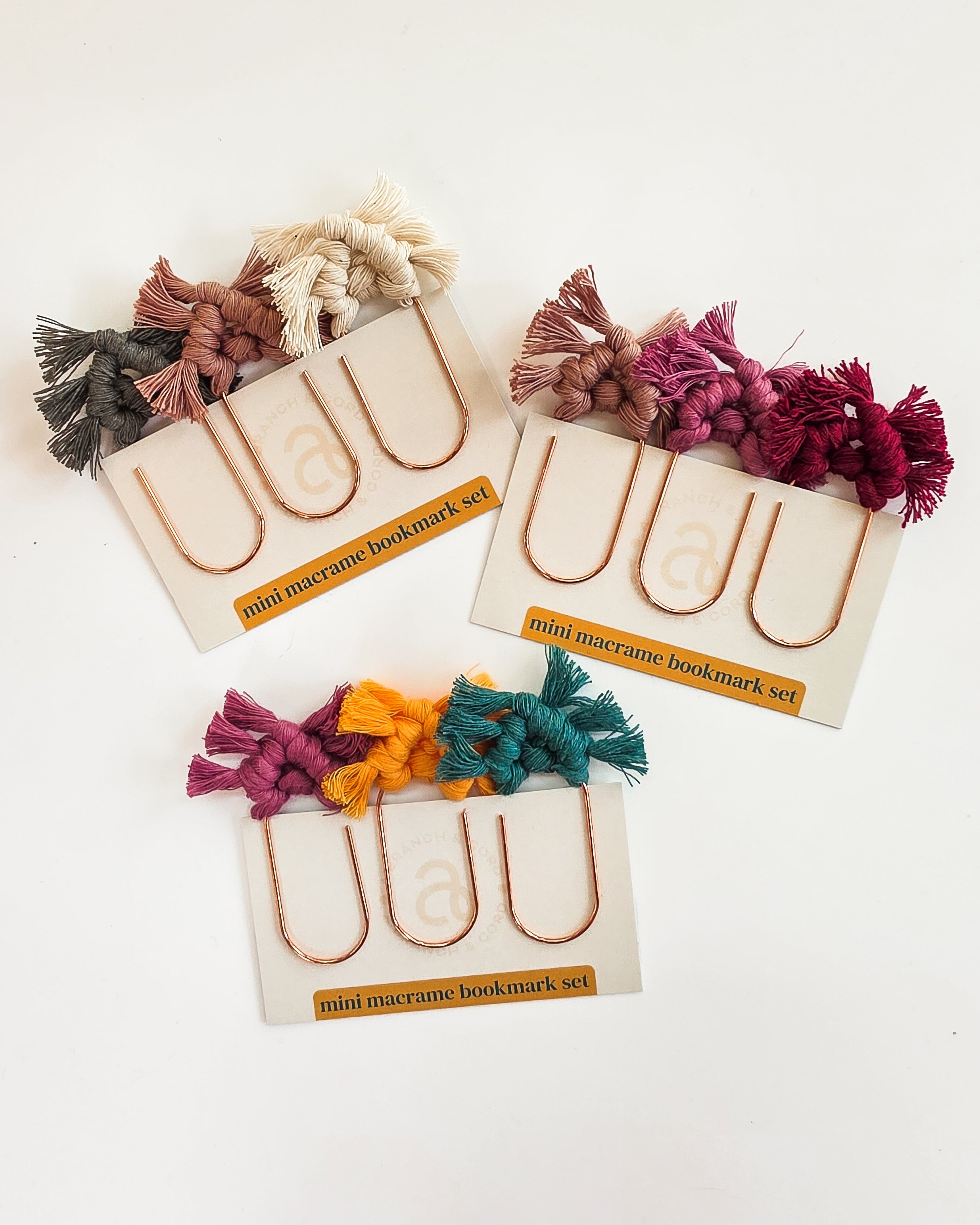 Three hand knotted mini macrame bookmarks in assorted jewel tone colors, resting on an open book