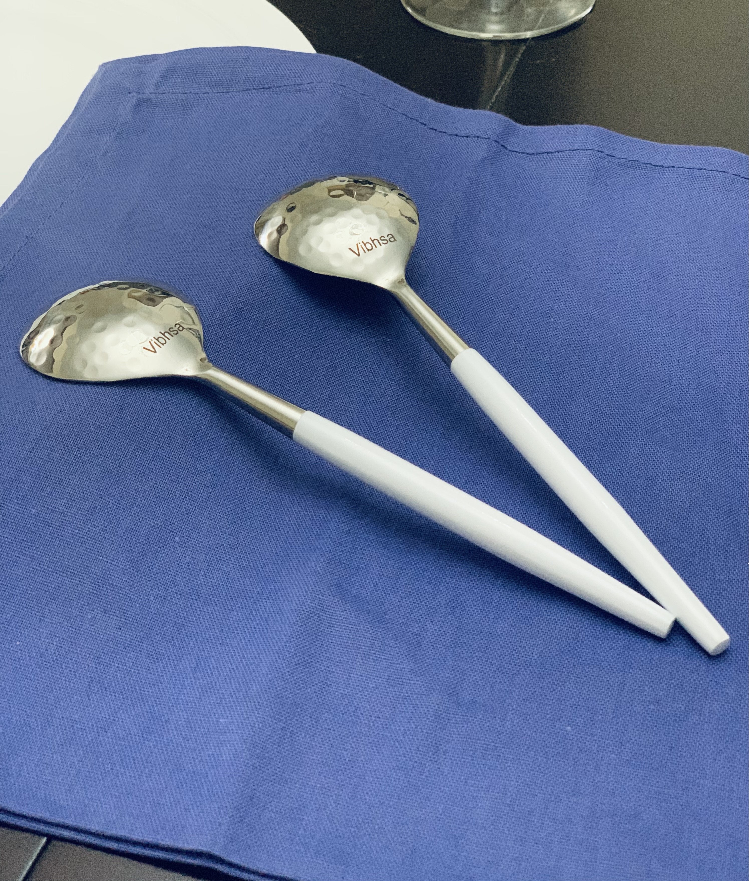 Vibhsa 6-piece stainless steel soup spoon set with hammered mouths and white handles arranged on a cloth background