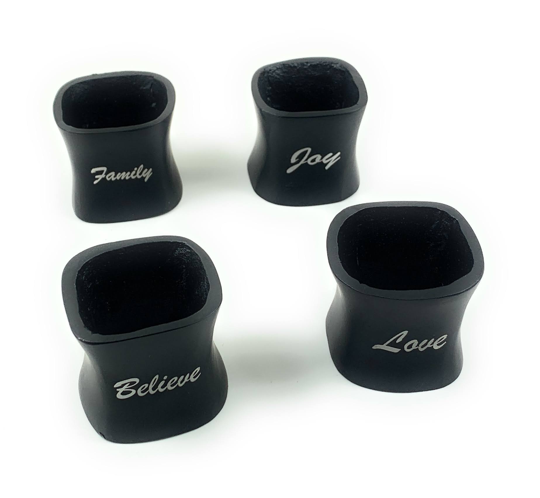 Four matte-finish black aluminum Christmas napkin rings engraved with Love, Believe, Joy, Family, arranged on folded white napkins