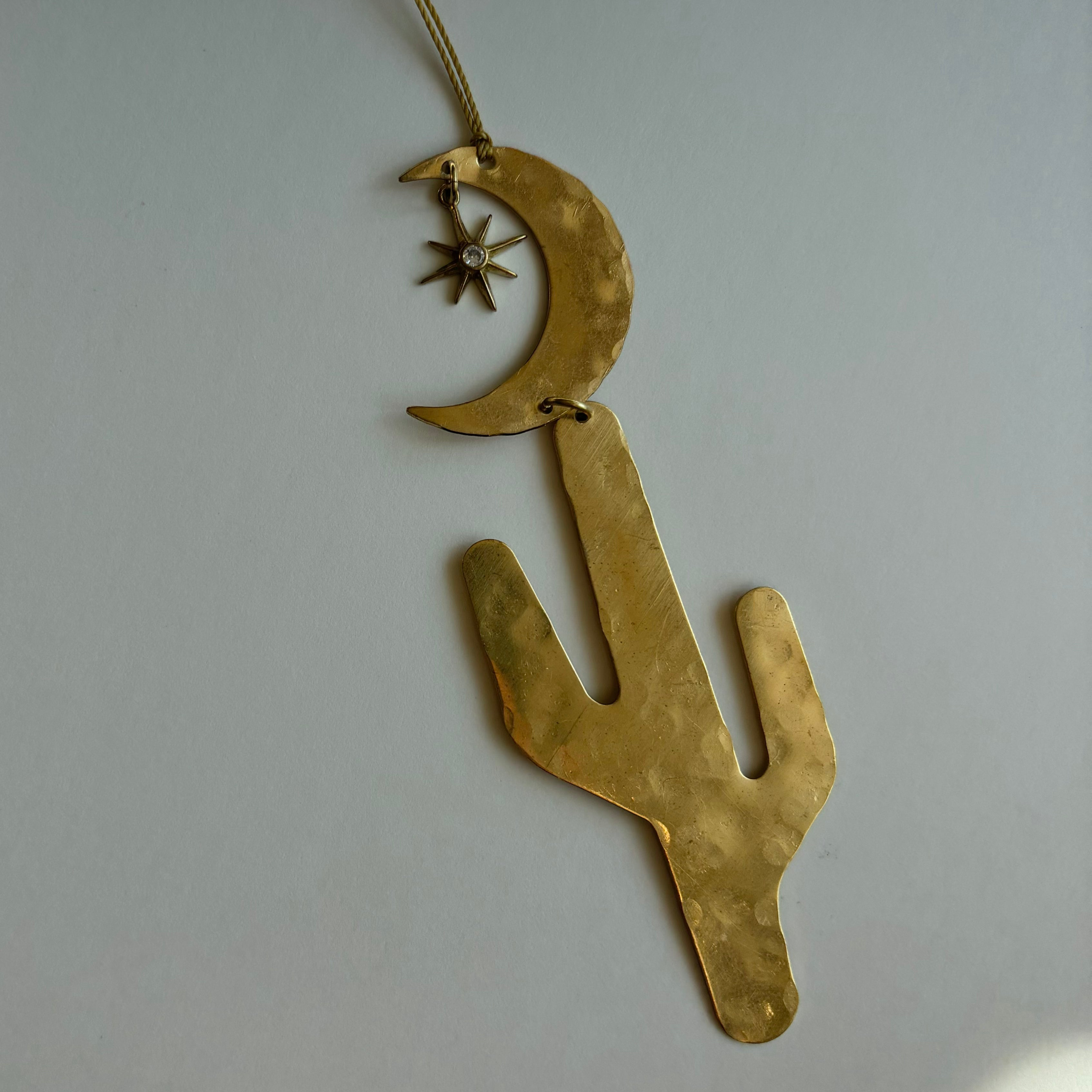 Desert Sky Ornament by Vida + Luz hanging on wall with ambient light highlighting hammered brass and crystal charm