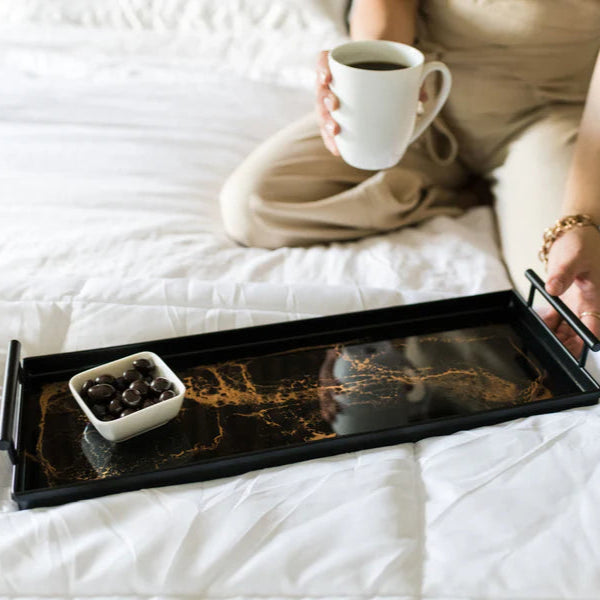 Person sitting on a bed holding a cup with a resin tray of coffee and cookies.