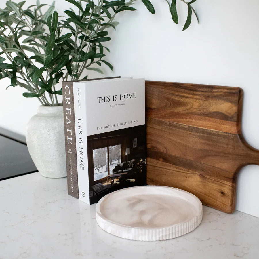 Open book titled 'This is Home' on a marble surface with a wooden cutting board and plant.