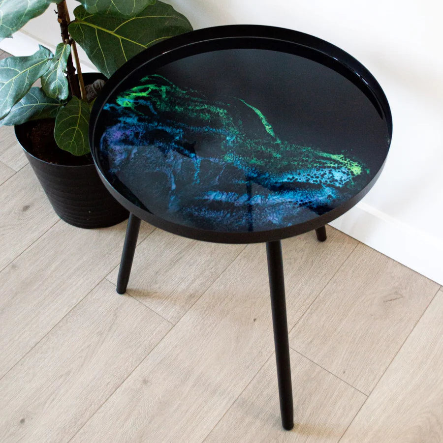 Black round resin table with a glossy surface next to a potted plant on a light wooden floor.