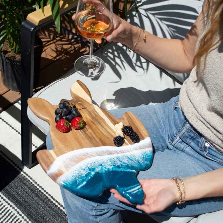 Person holding a glass of wine with a resin charcuterie board and fruits, sitting outdoors.