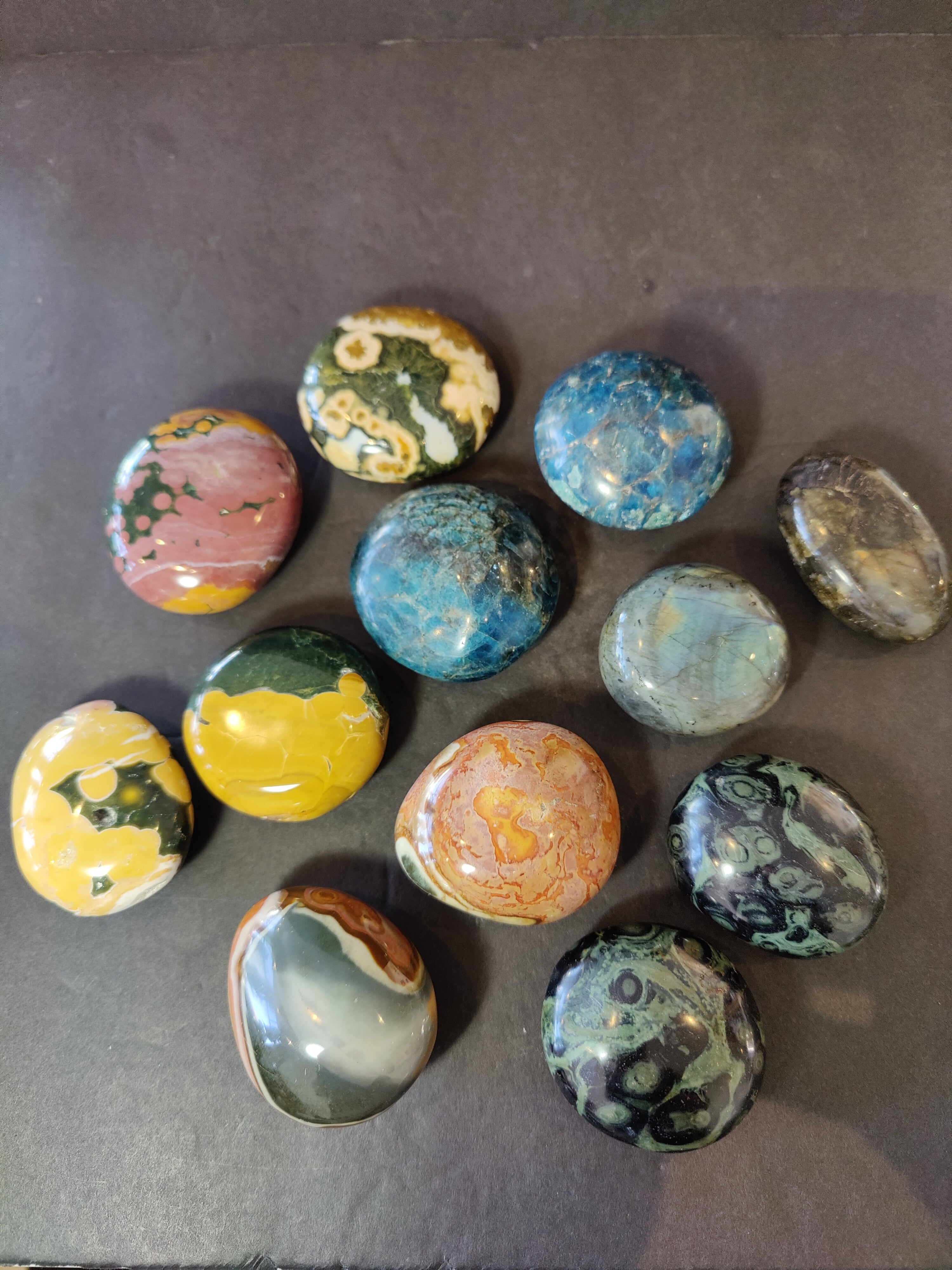 Assorted polished palmstones including Ocean Jasper, Blue Apatite, Kambaba Jasper, Polychrome Jasper, and Labradorite stones arranged on a wooden surface