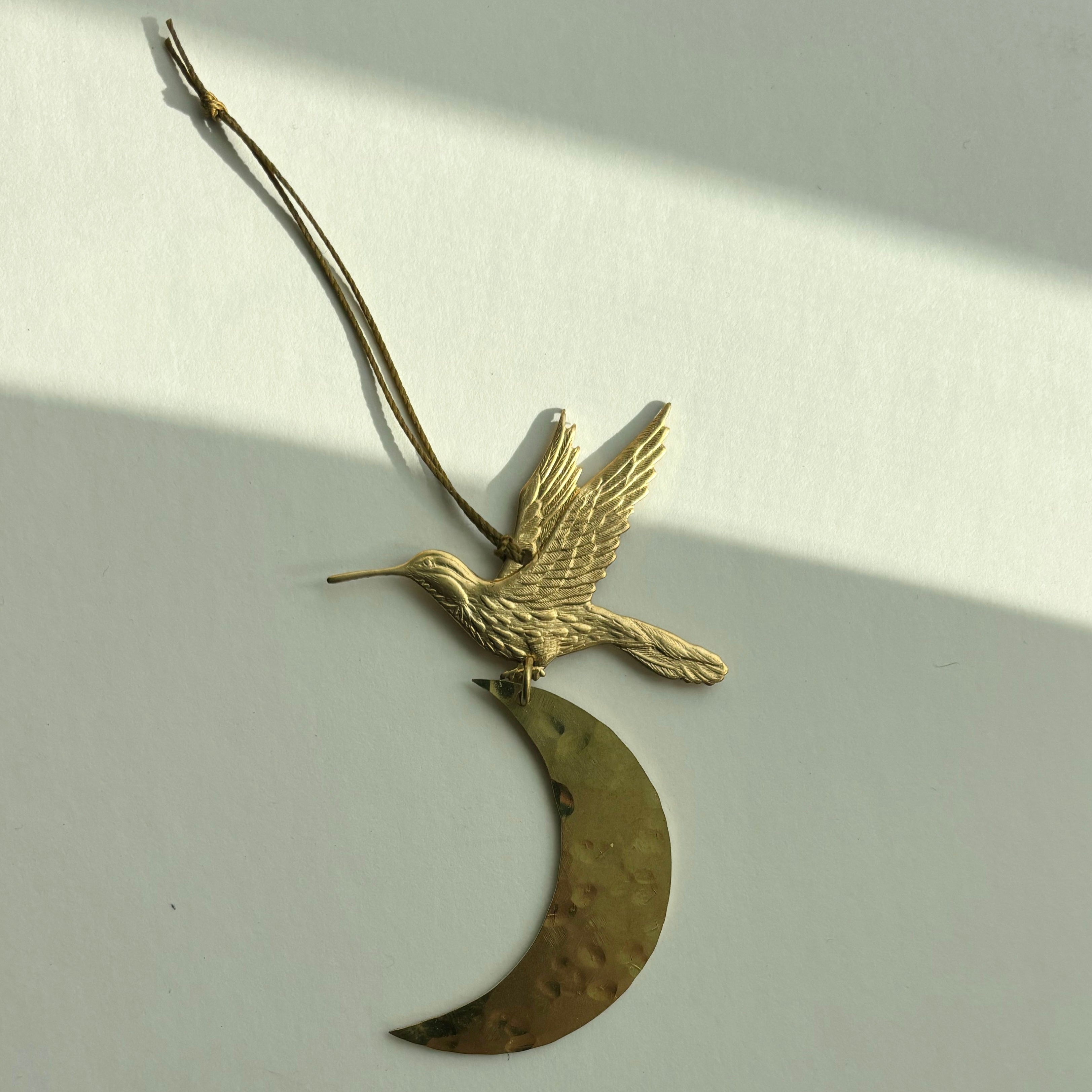 Over the Moon Ornament by Vida + Luz hanging against a white wall, showing slender crescent shape in hammered brass.