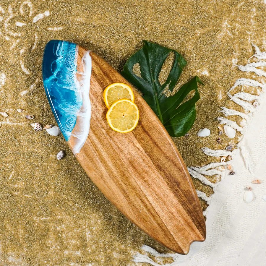 Wooden surfboard-shaped charcuterie board with blue resin accent, holding cheese and fruit on a patio table