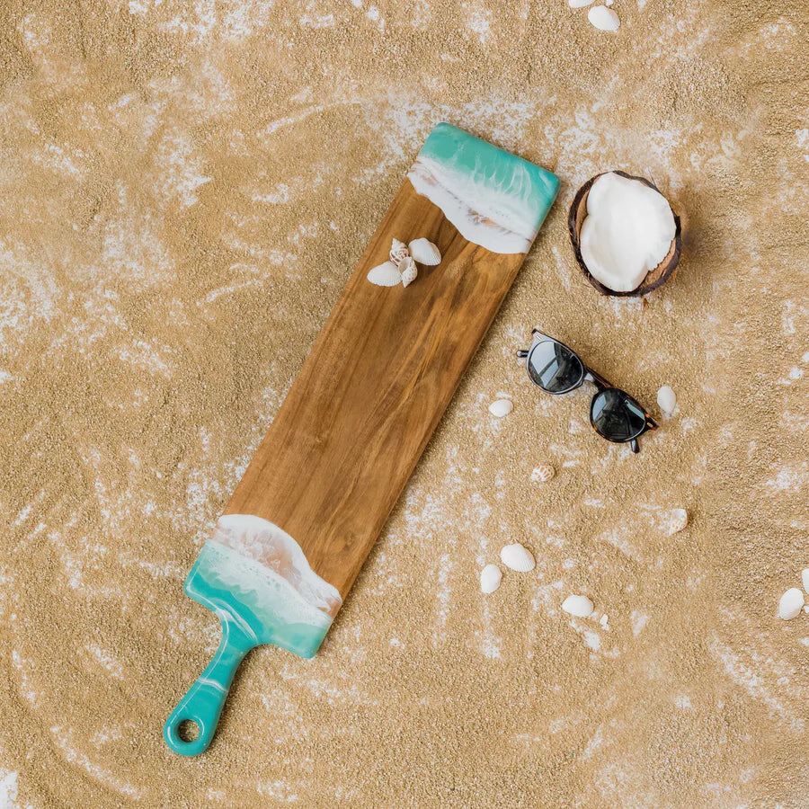 Wooden baguette resin serving tray with teal handle and white foam design on sandy surface with coconut and sunglasses.