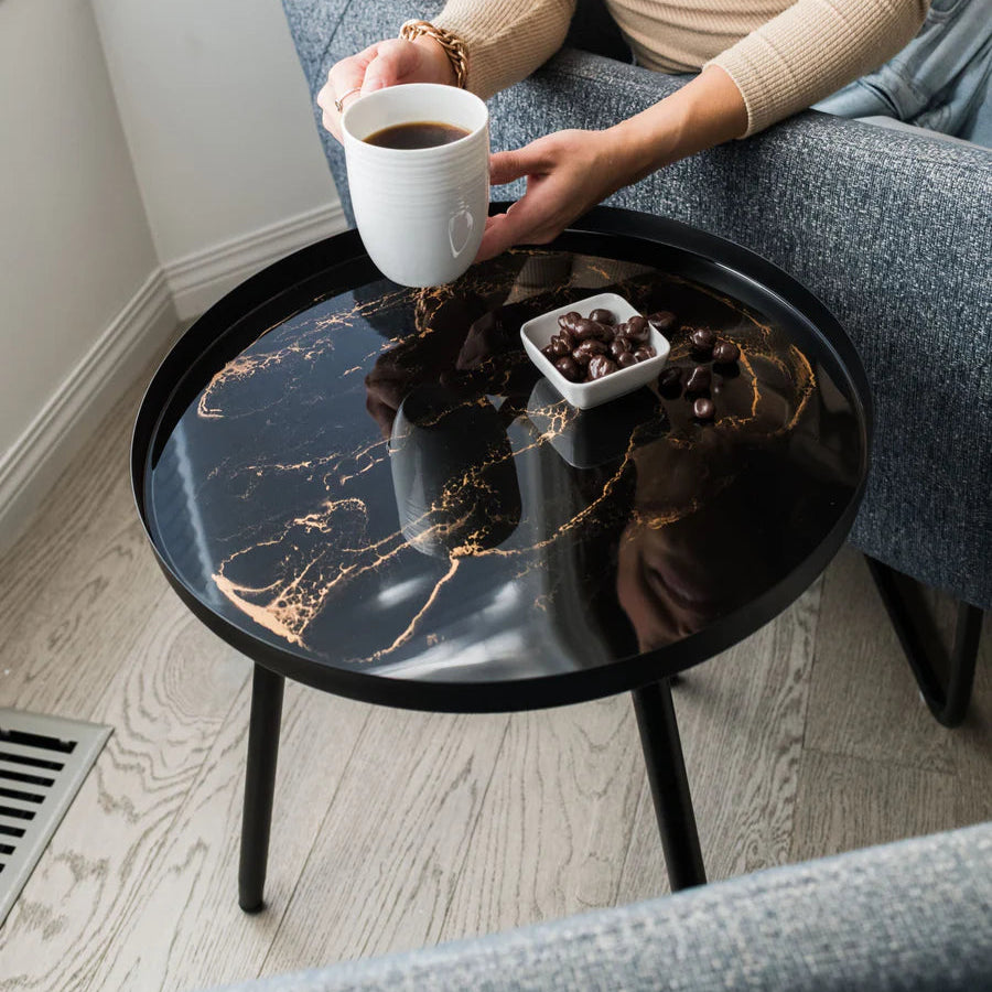 Person sitting on a gray sofa holding a white mug with coffee, next to a black round small resin side table.