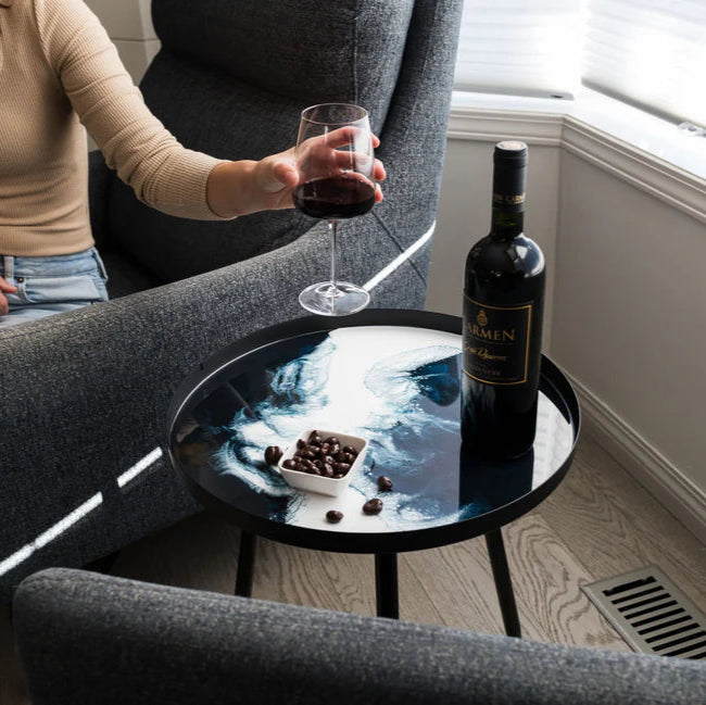 Person sitting on a couch holding a glass of wine with a bottle and snacks on a small resin table.