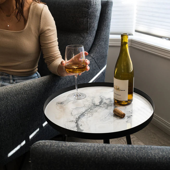 Person holding a glass of wine next to a bottle on a marble resin table in a cozy room.