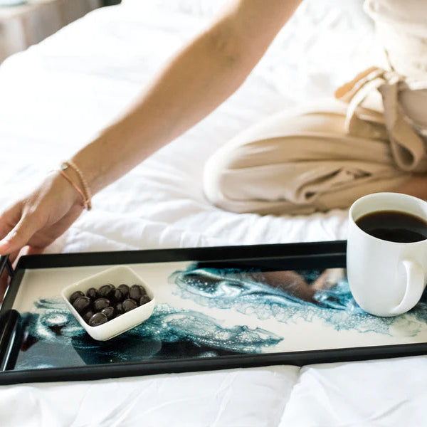 Person holding a resin tray with a cup of coffee and a bowl of blackberries on a bed.