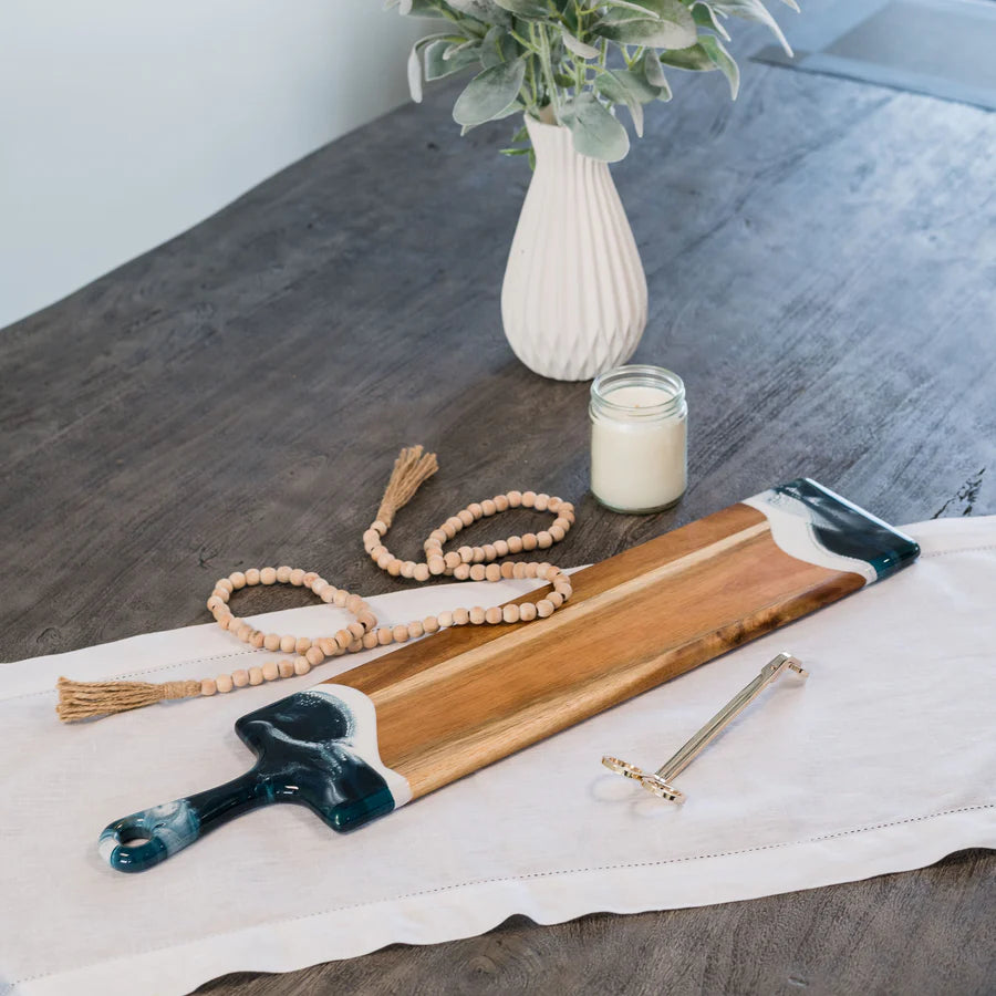 Wooden baguette resin serving tray with decorative handles on a table with a candle and vase in the background.