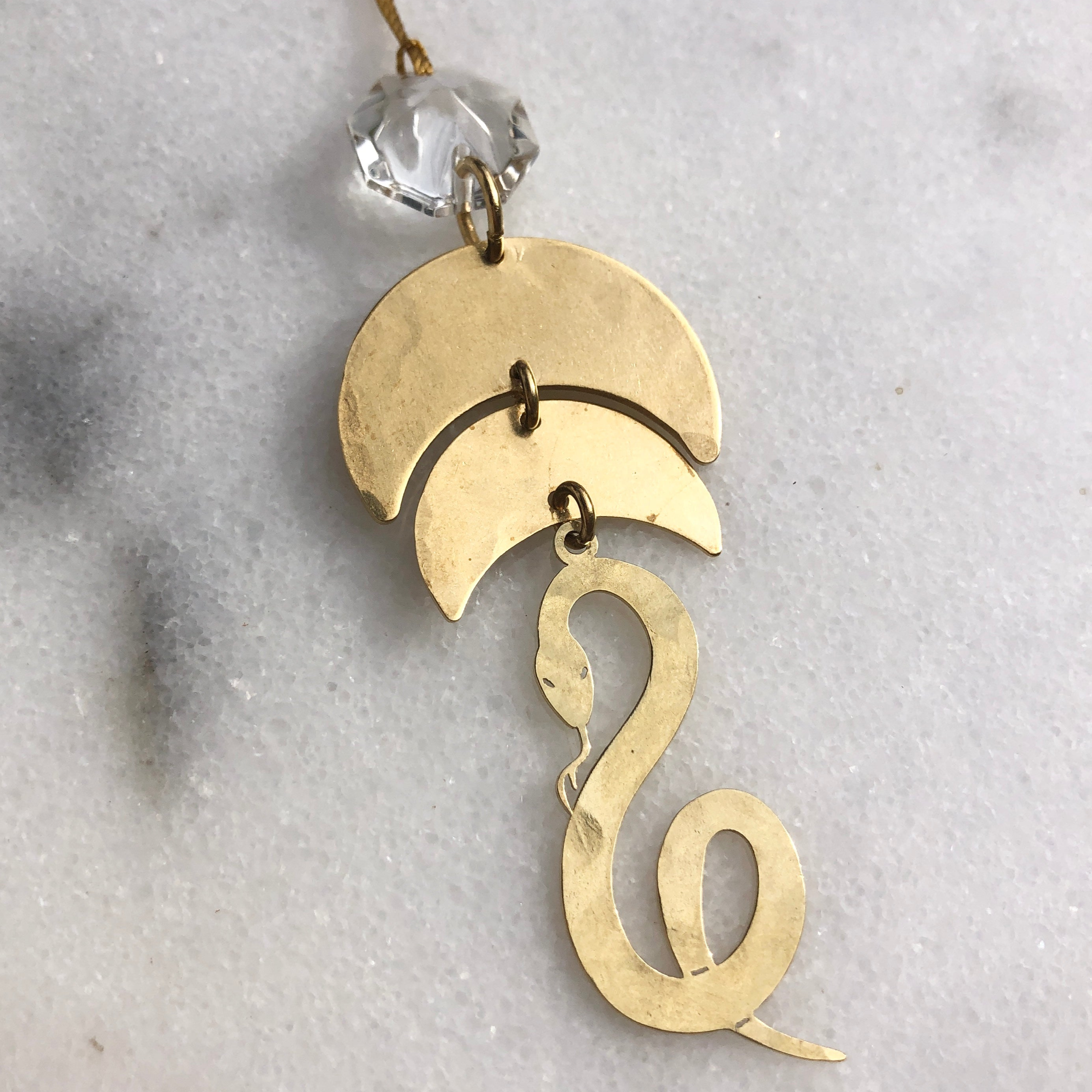 Serpentine Ornament featuring hammered brass and a vintage glass crystal, hanging with cord on a white background