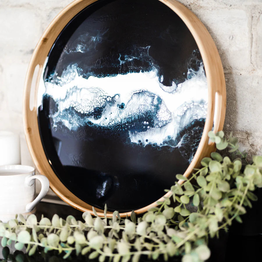 Round decorative resin serving tray with black and white abstract design on a wooden rim, placed on a surface with greenery.