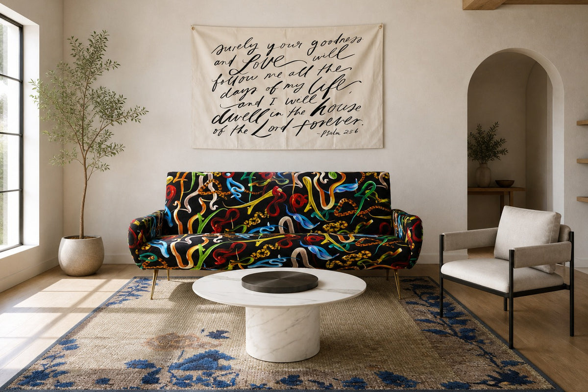 Living room with colorful snake-patterned sofa centered beneath a beige wall hanging with script, a round white marble coffee table, neutral armchair, woven rug, and a potted plant by a large window.