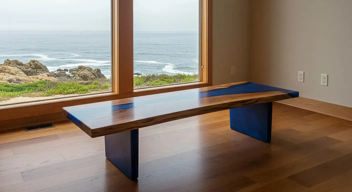 A low dining bench with a wooden top featuring natural wood grain and navy blue resin accents on the edges, supported by two navy blue resin legs, set on a light wooden floor near a large window with an ocean and rocky shoreline view.