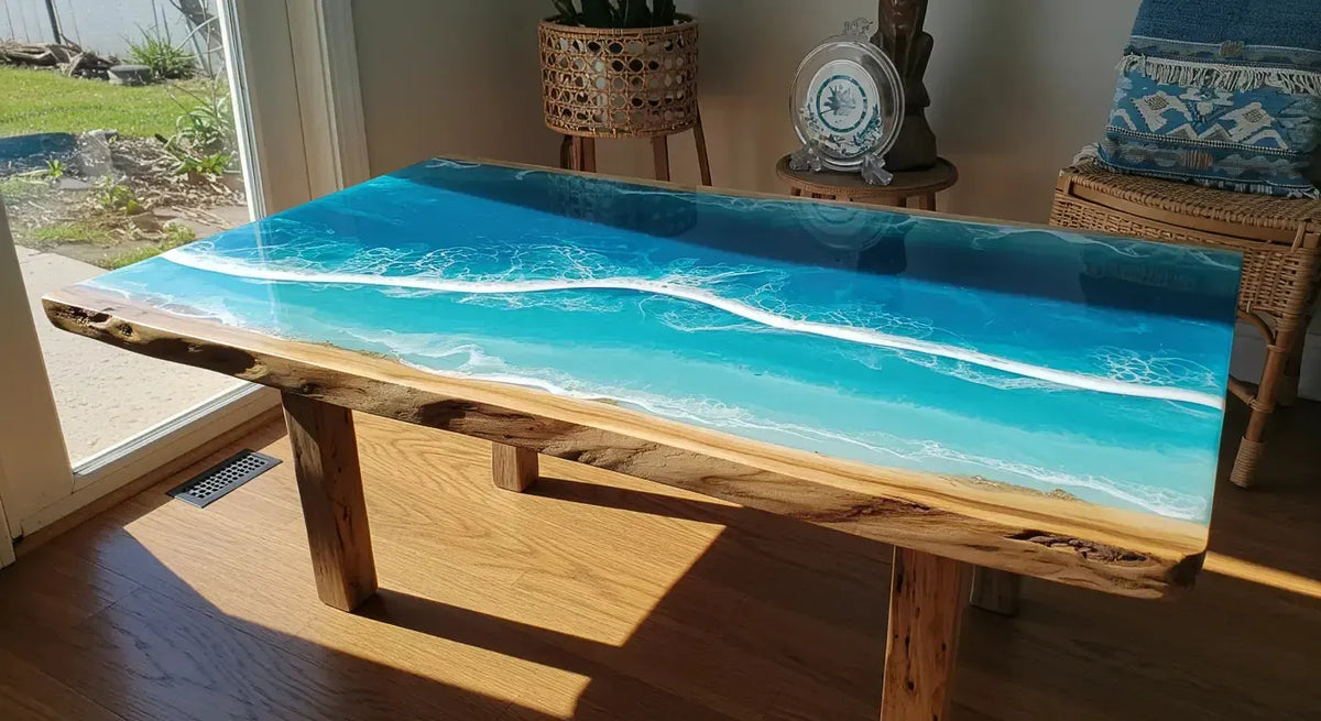 Live-edge wooden table with a turquoise ocean-inspired resin surface and white wave details beside a sunlit window.
