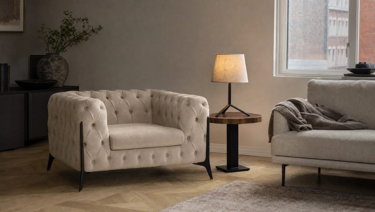 A cozy living room corner featuring a beige tufted armchair with black legs, a round wooden side table with a lit lamp, a nearby beige sofa draped with a gray throw, and a large window letting in soft natural light.