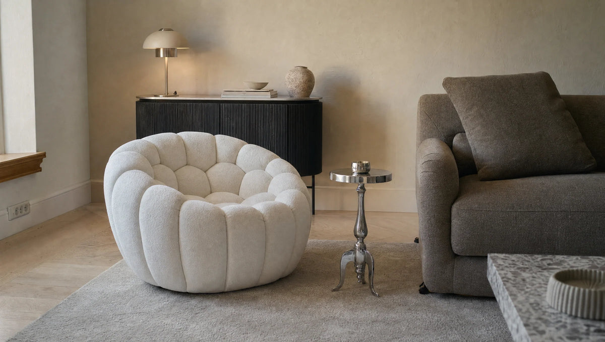 A neutral-toned living room corner features a rounded, tufted beige armchair, a small ornate silver side table, a dark gray sofa with a cushion, and a black sideboard with a lamp and decorative items.