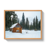 Small rustic wooden hut with smoke rising from a chimney, surrounded by snow and tall evergreen trees in a foggy winter landscape.