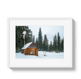 Rustic wooden hut with smoke rising from its chimney, surrounded by snow and tall evergreen trees on a foggy winter day