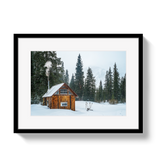 Rustic wooden warming hut with metal roof emitting smoke from chimney, surrounded by snow and tall evergreen trees in a misty forest clearing.