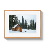 Rustic wooden cabin with smoke rising from the chimney, surrounded by snow and tall evergreen trees under a pale sky.