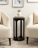 Black round wood drink table with curved vertical slats and a bottom shelf, placed between two cream upholstered armchairs on a patterned rug