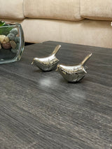 Two small silver bird figurines with textured feather details placed on a dark wood surface near a glass container with multicolored stones and a beige couch background.