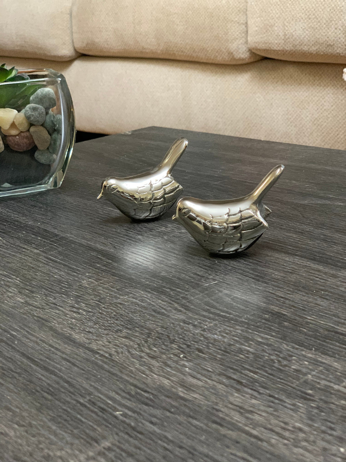 Two small silver bird figurines with textured feather details placed on a dark wood surface near a glass container with multicolored stones and a beige couch background.