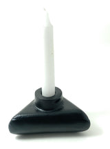 Black triangular candle holder with rounded edges holding a tall white taper candle, shown against a white background with soft shadows.