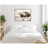 Sepia-toned framed canvas print of two Texas Longhorn cattle with large curved horns standing in a pasture, hung on a white wall above a white bed with pillows.
