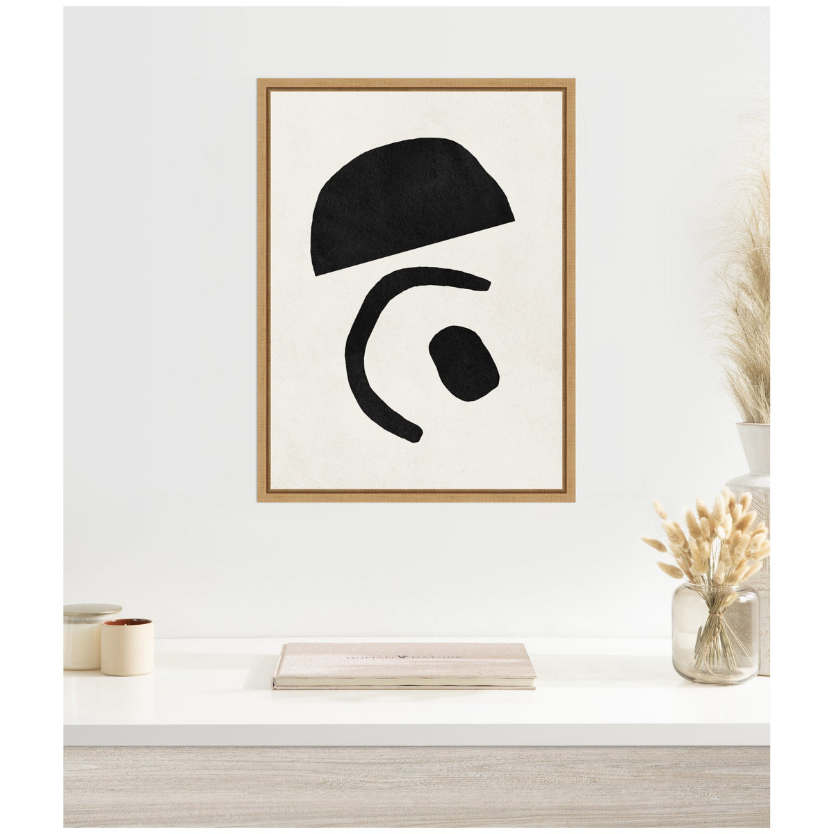 Framed abstract canvas with three irregular black shapes on white background in a natural wood frame, hanging above a white desk with neutral decorative items.