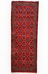 Long red vintage Mashhad wool runner with repeating floral and geometric motifs in blue, white, and brown, framed by multiple decorative borders.