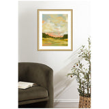 Impressionistic landscape painting with soft clouds in blue and peach above a green and orange field, framed in natural wood and hung above a dark green armchair and potted olive tree.