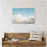 Soft white clouds against a light blue sky on a framed canvas above a rustic wooden bench with an open book and a beige pillow reading 'welcome home'.