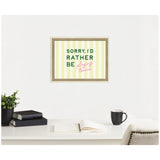 Framed wall art with green and pink text saying 'SORRY, I'D RATHER BE playing tennis' on a yellow and white striped background, hanging above a white desk with books, a plant, an open notebook, and a black coffee mug.