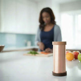 Copper cylindrical Qi-Shield by Waveguard with wooden top and base on white kitchen counter, blurred person chopping vegetables in background.