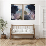 Two framed abstract canvas paintings with dark navy, pale pink, gray, and olive brushstrokes above a wooden bench with white cushions set against a white wall