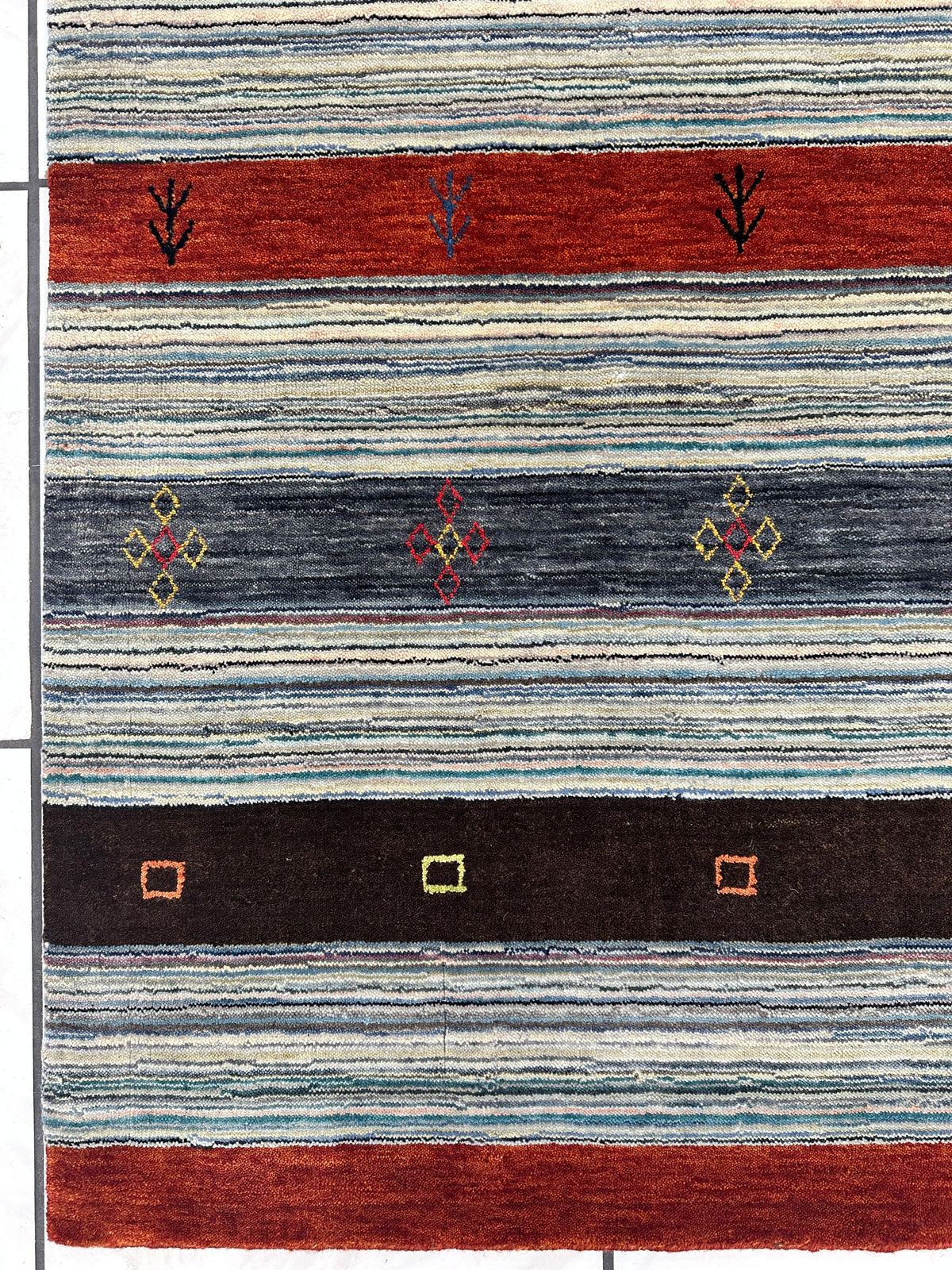 Striped woven rug with alternating bands of red, gray, and black featuring small geometric and floral motifs in yellow, red, and blue on a textured white and blue background.