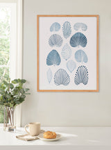 Framed print with various blue and teal stylized coral shapes arranged in a grid on a white background, hanging on a pale wall above a table with a plant, cup, and croissant.