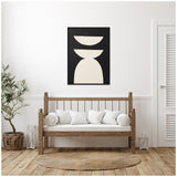 Black framed abstract canvas with three large white geometric shapes, hung above a wooden bench with white cushions in a minimalist room with light walls and natural wood flooring.