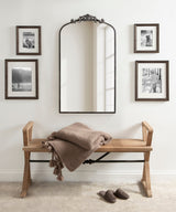 Arched mirror with ornate dark metal frame hanging above a wooden bench with a folded beige knit blanket, flanked by four black-and-white framed photographs on a white wall.