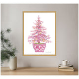 Framed canvas artwork of a pink Christmas tree decorated with multicolored ornaments in a detailed pink floral-patterned pot, hanging on a beige wall above a wooden shelf.