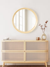 Round wall mirror with light wood frame hung above a mid-century modern wooden dresser featuring woven cane drawer fronts, styled with neutral decor items.