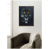 Framed art print showing a collection of nine varied butterflies with dark and neutral tones on a deep blue background, hanging above an olive green armchair with a white knit throw.