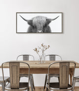 Framed black and white photograph of a Highland cow's head with long hair and wide horns, hung above a rustic wooden dining table with four metal chairs and a vase with dried flowers.
