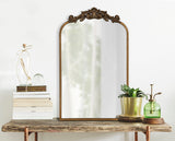 Traditional arched mirror with ornate dark bronze decorative crest on top, standing on rustic wooden table alongside stacked white books, potted plant, and green glass mister.