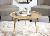 Round gold coffee table with a scalloped edge, slender tapered legs, topped with a small white flower arrangement and books, set on a fluffy white rug.